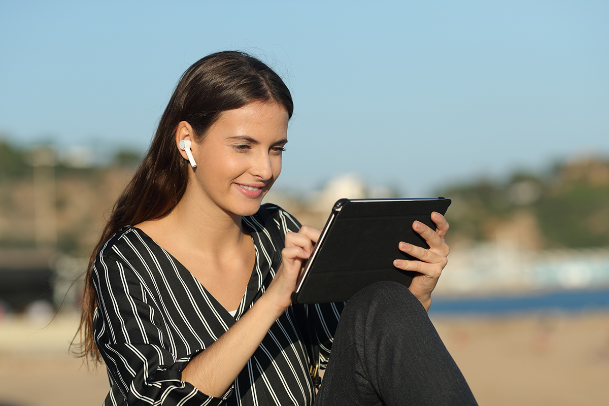 Случайная счастливая женщина электронного обучения с планшетом и беспроводными наушниками, сидящая на пляже