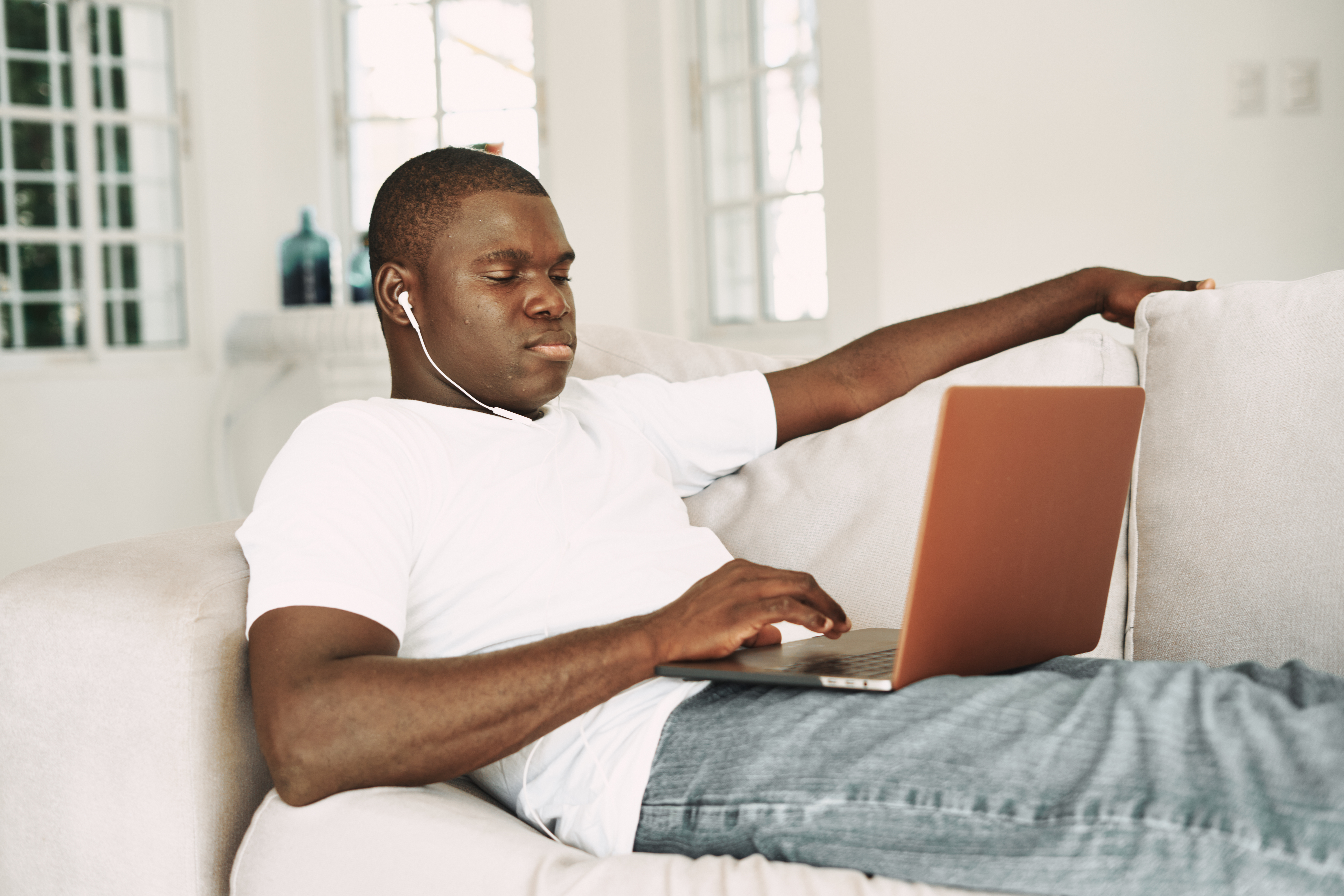 A nice guy of African appearance lies on the couch with headphones and a laptop on his lap