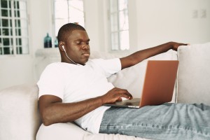 Simpatičan momak afričkog izgleda leži na kauču sa slušalicama i laptopom