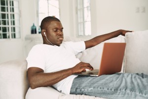 A nice guy of African appearance lies on the couch with headphones and a laptop